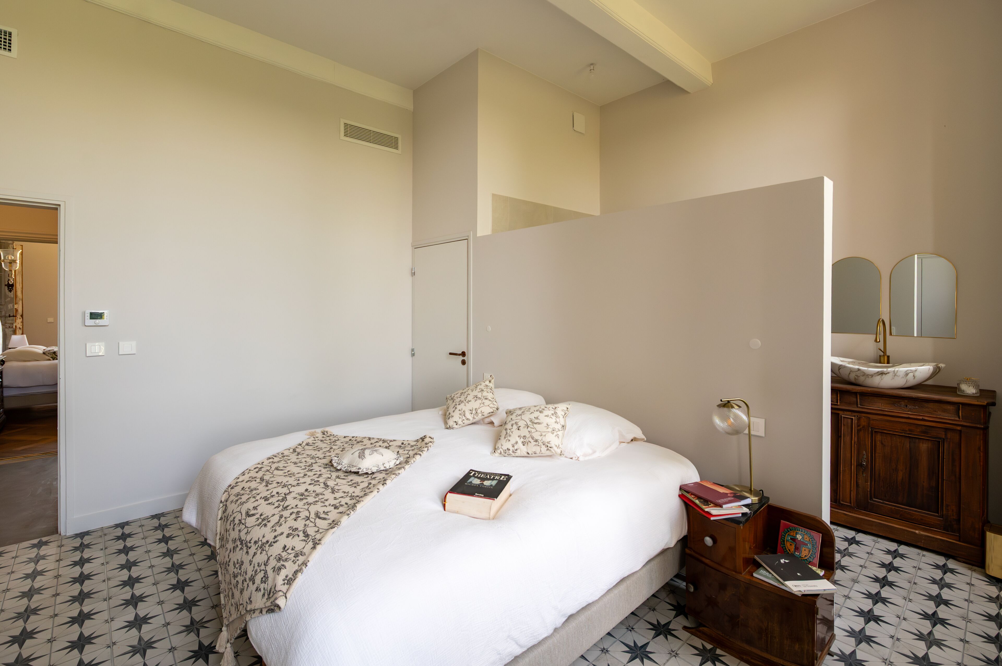 A bedroom with a bed, a bedside table, a mirror, and a bathroom with a sink.