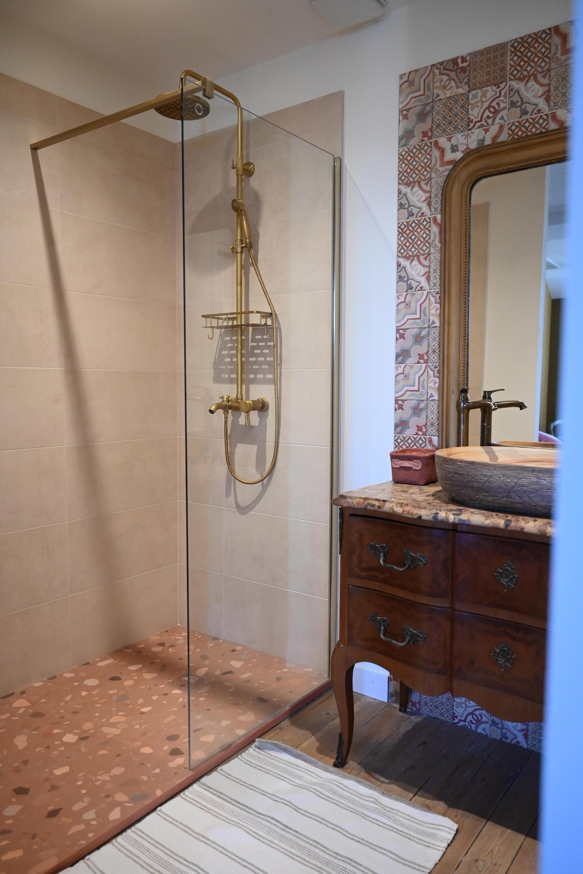 Ducha con puerta de vidrio, tocador de madera con espejo y salpicadero de azulejos estampados.