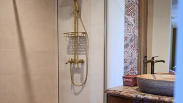 Ducha con puerta de vidrio, tocador de madera con espejo y salpicadero de azulejos estampados.