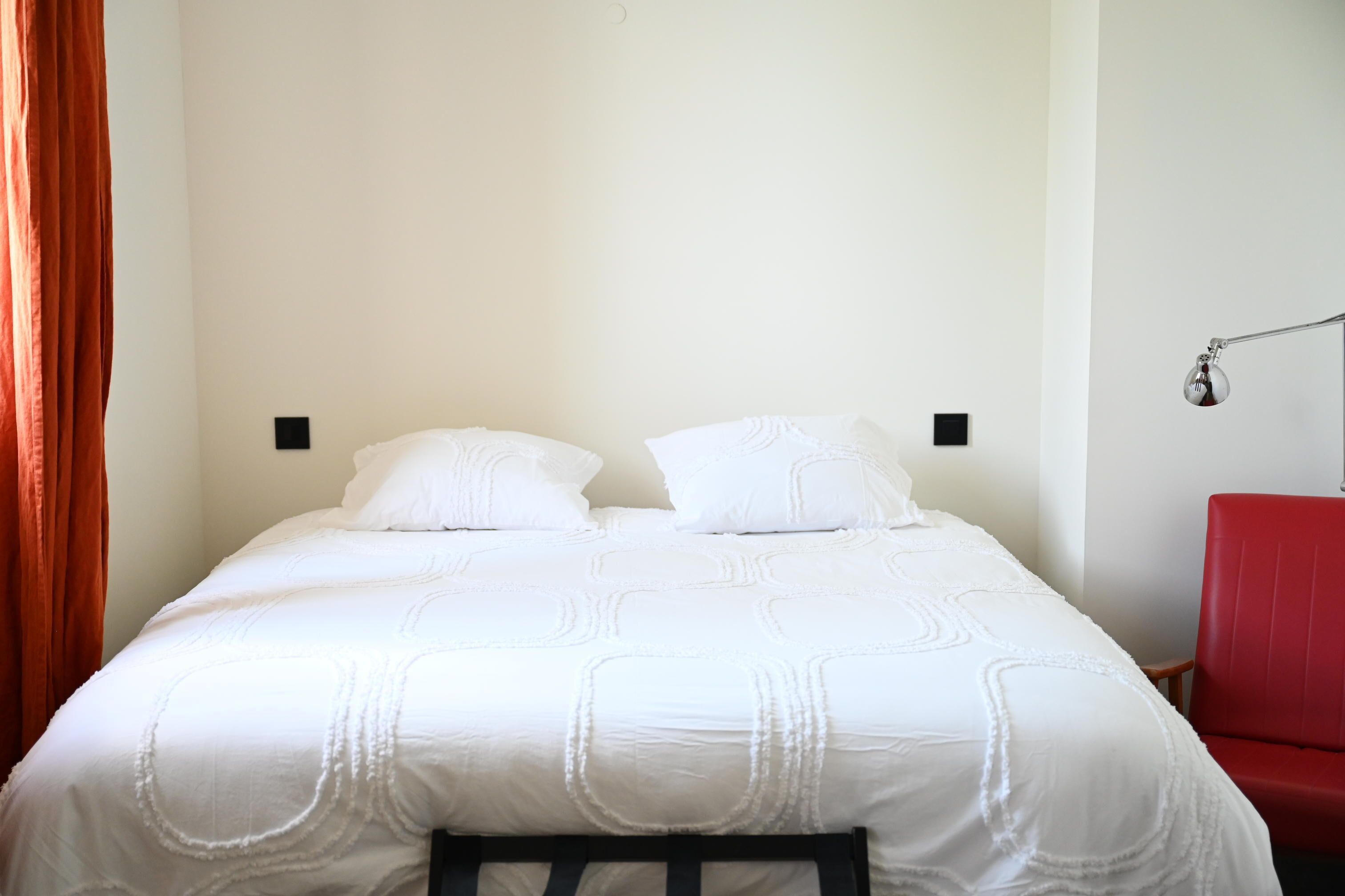 Una cama con ropa de cama blanca, una silla roja y una lámpara de pared.