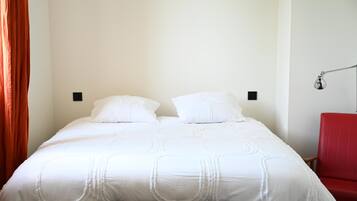Una cama con ropa de cama blanca, una silla roja y una lámpara de pared.