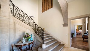 A grand staircase with ornate black iron railings and a marble floor. A small table with decorative items is placed at the base. - Château de Scalibert (Teyssode)