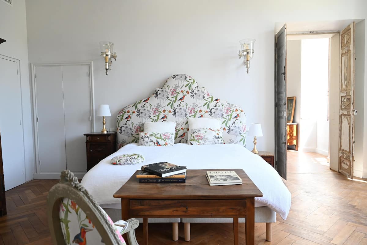 A bedroom with a floral headboard, a wooden bedside table with books, and a white bedspread.