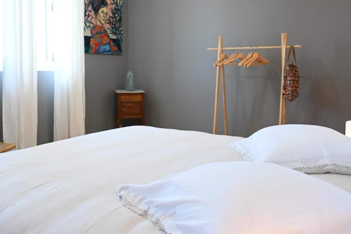 A bedroom with a large bed, a wooden drying rack, a small bedside table, and a painting on the wall.