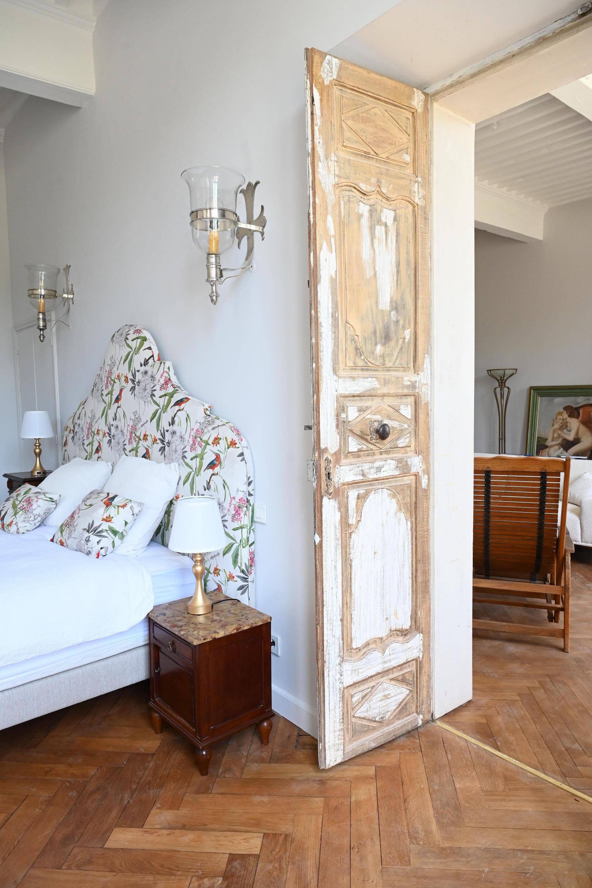 A bedroom with a floral headboard, a wooden nightstand, a bed with white linens, and a large open door leading to another room.