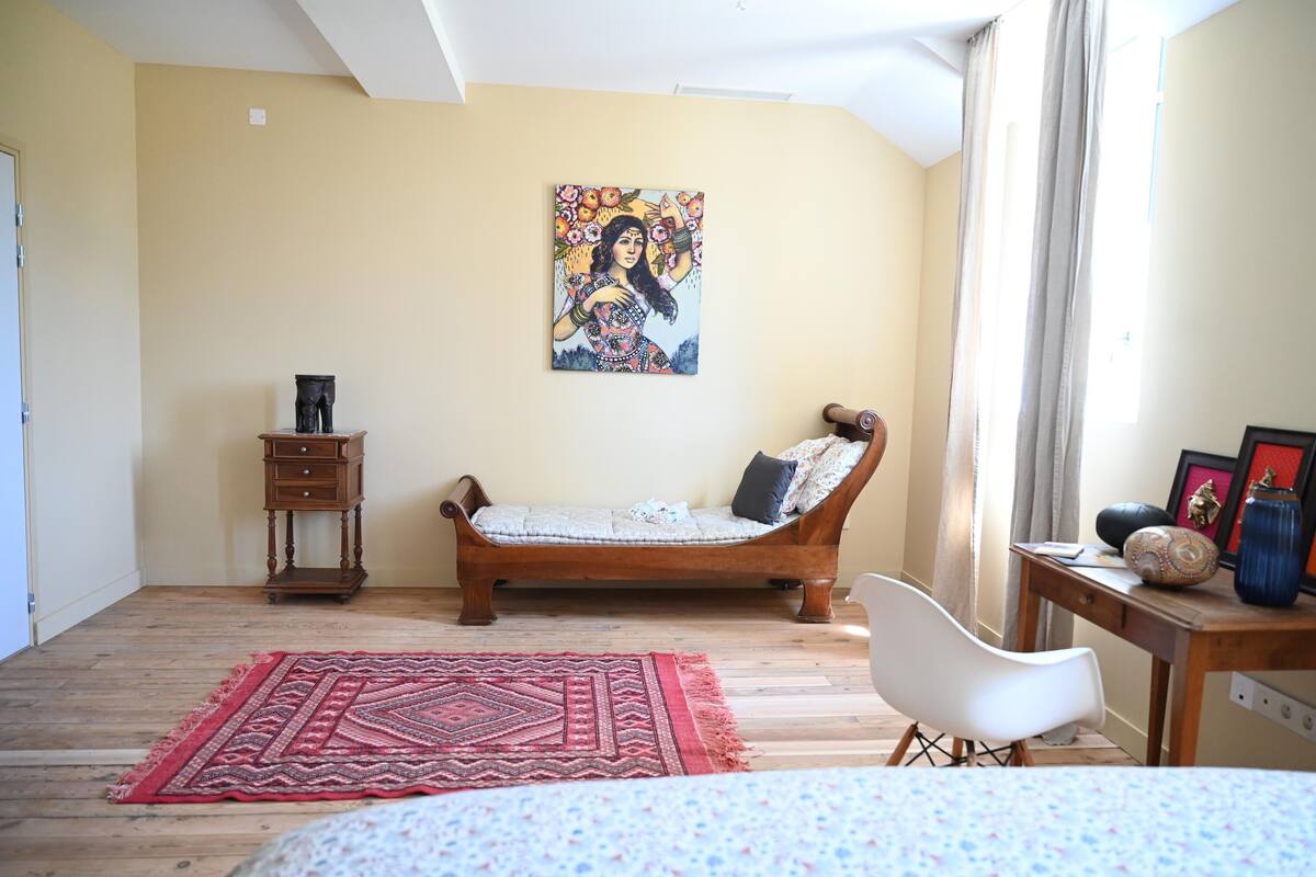 A bedroom with a wooden bed, a red patterned rug, a white chair, a wooden nightstand, and a painting on the wall.