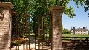 A gated entrance with a brick pillar and a metal gate, leading to a large building with a brick facade and multiple windows. - Château de Scalibert (Teyssode)