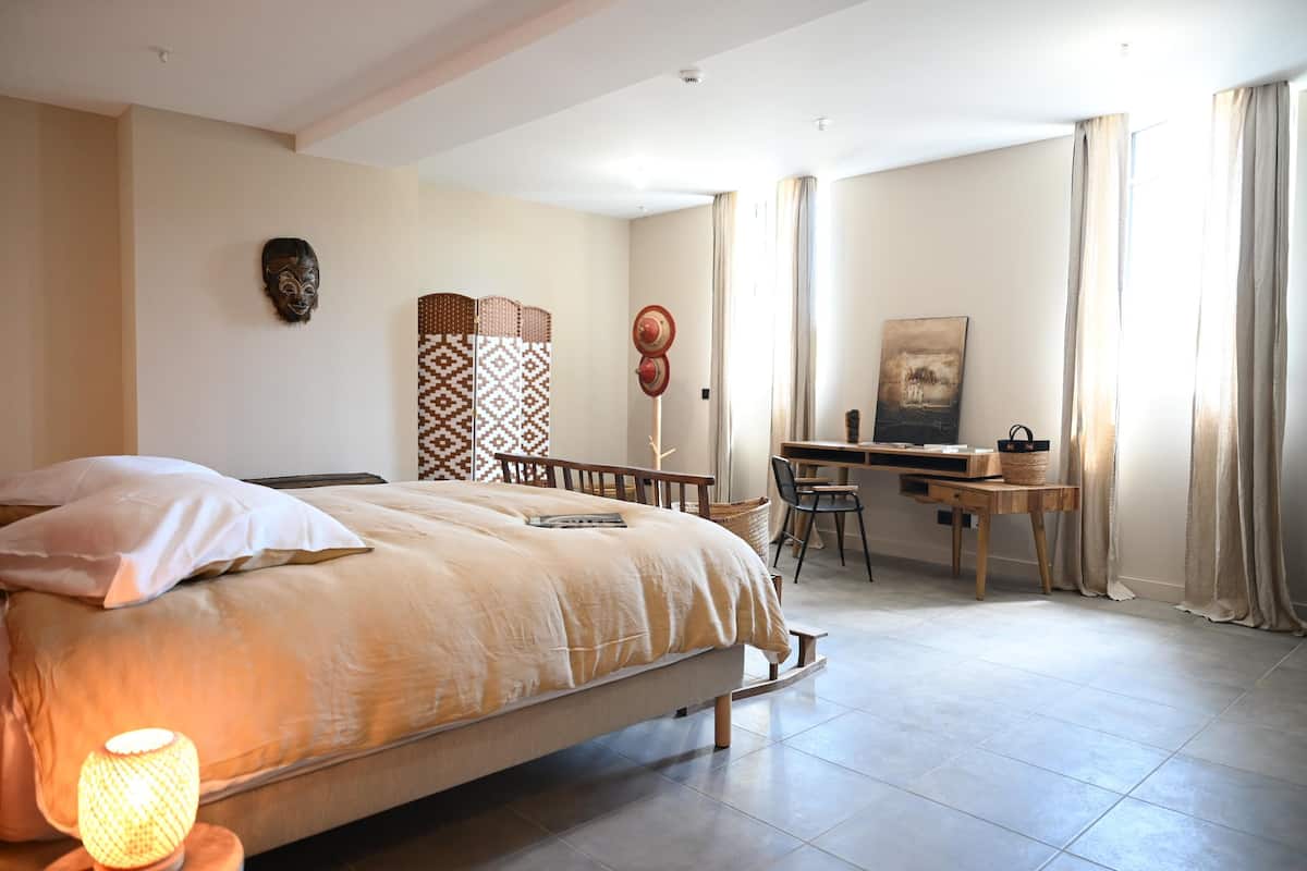 A bedroom with a large bed, a desk, and a chair. There is a wall-mounted shelf with baskets and a decorative mask.