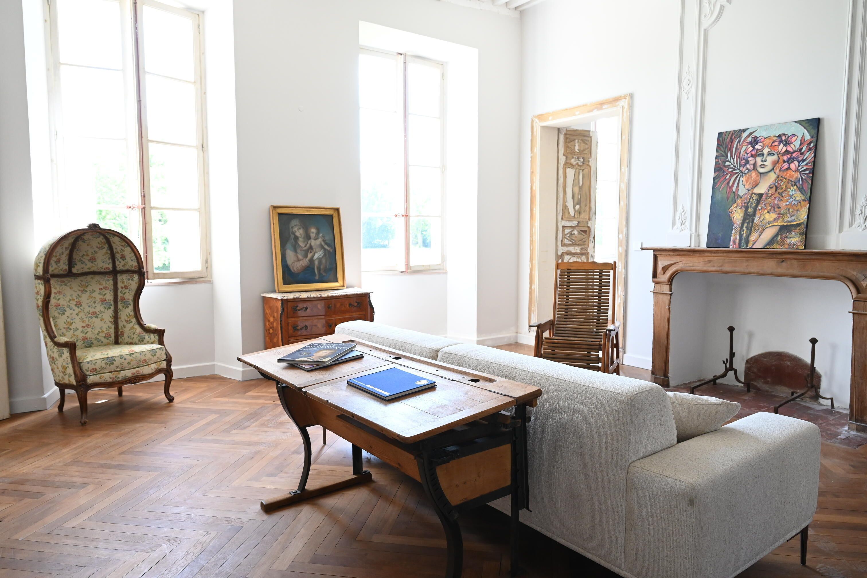 A room with a wooden floor, a sofa, a chair, a table with a book, a fireplace, and a painting on the wall.