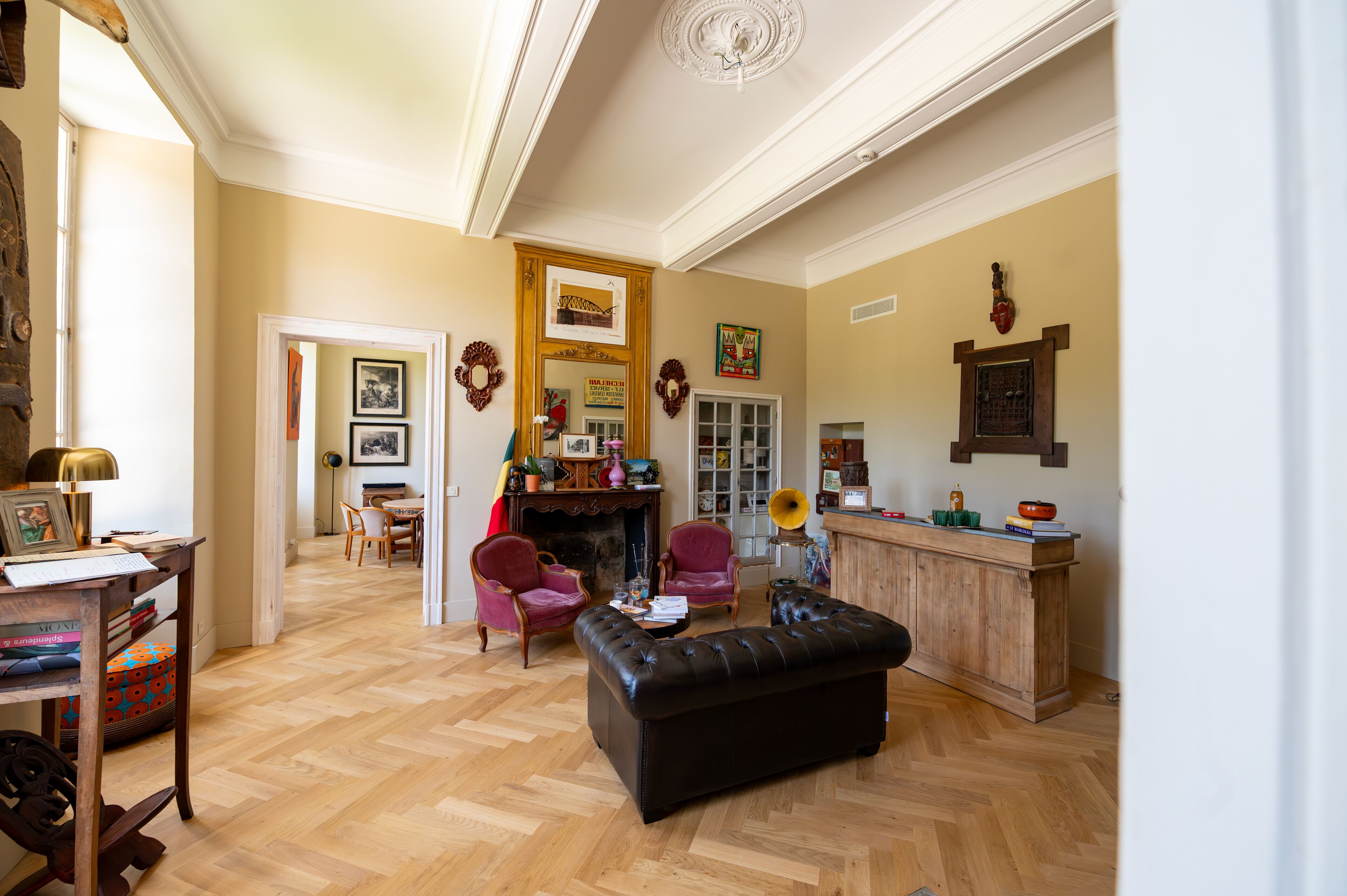 Ampla sala de estar com piso de madeira, um banquinho de couro preto e uma lareira. Há duas poltronas vermelhas e uma mesa de centro de madeira.