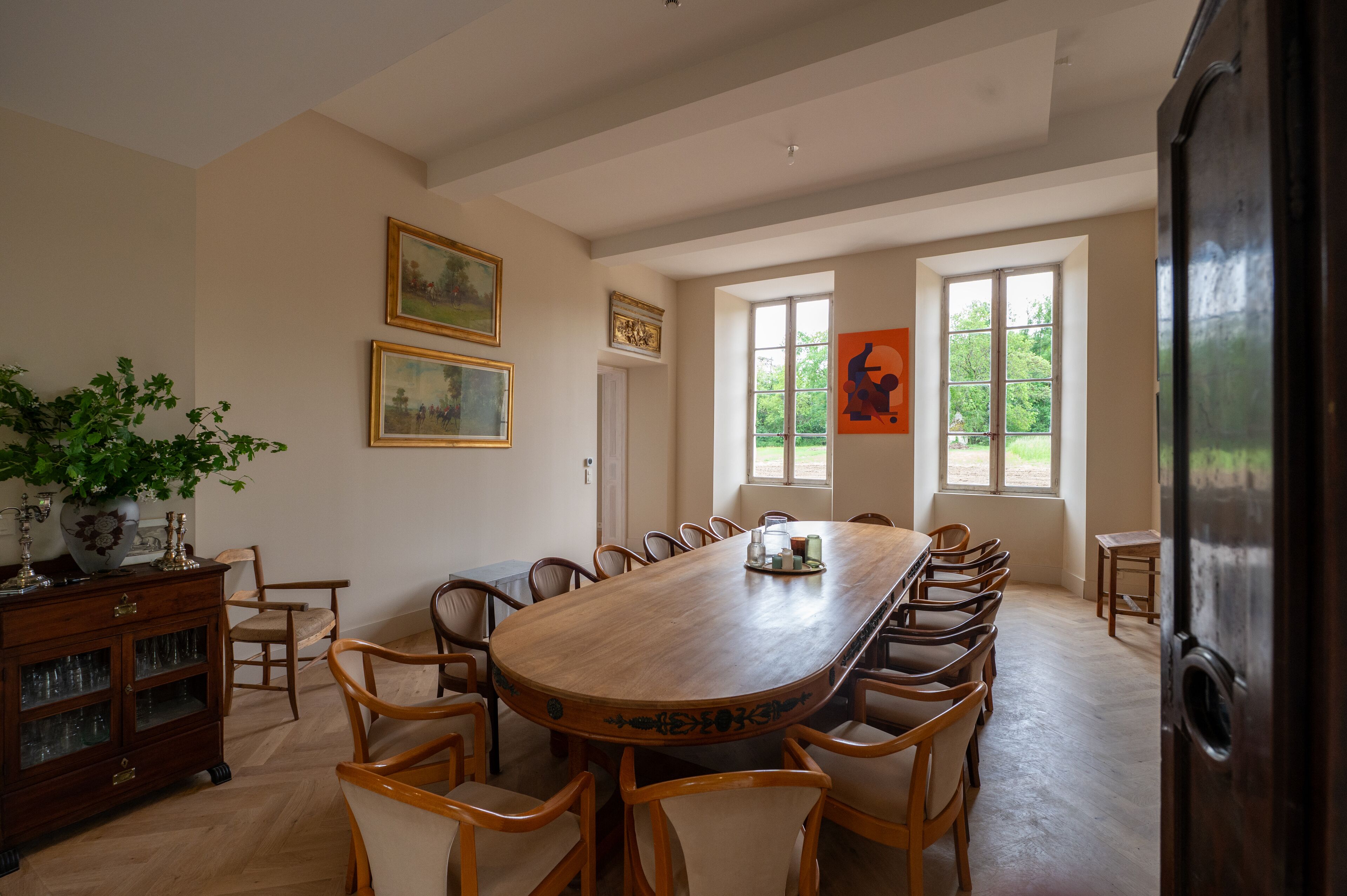 Comedor con una larga mesa de madera rodeada de sillas, un aparador con puertas de vidrio y obras de arte enmarcadas en las paredes.