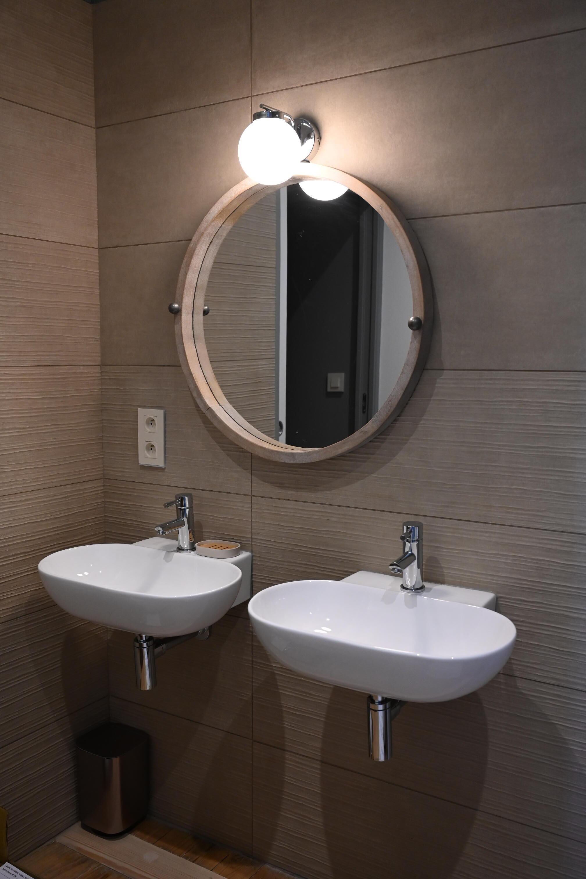 Een badkamer met twee witte wasbakken, een ronde spiegel met verlichting erboven en een prullenbak.