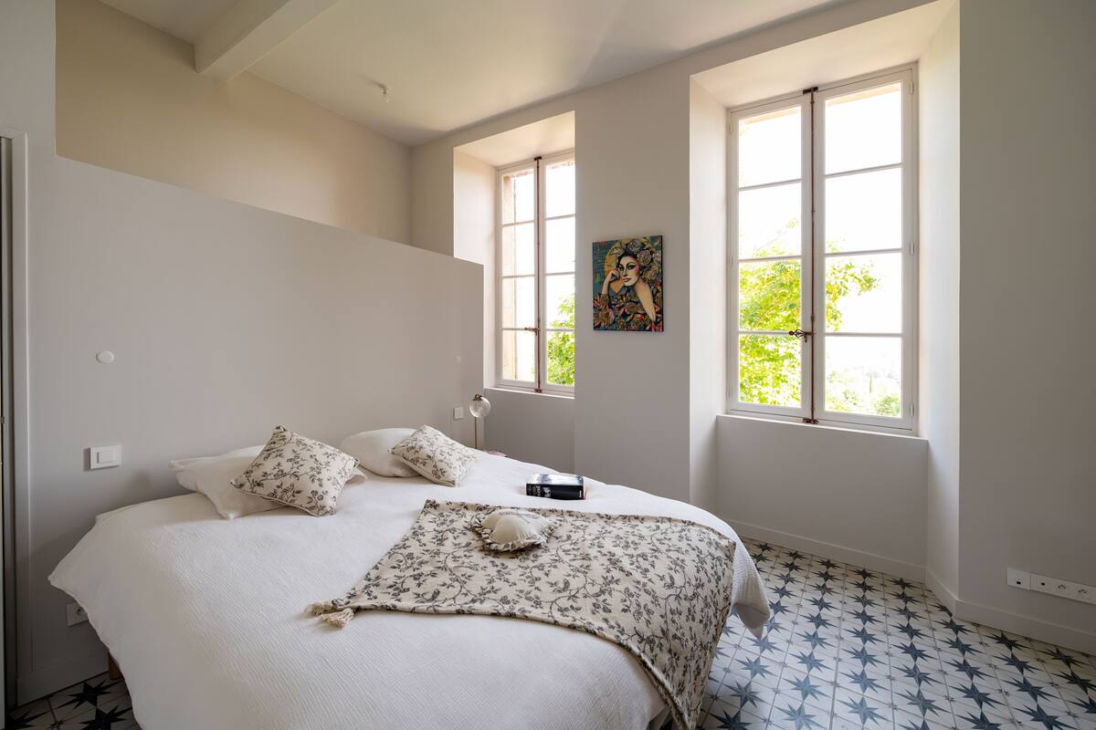 A bedroom with a bed, two windows, a painting, and a patterned floor.