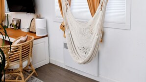 Ein Zimmer mit Holzboden, weißer Wand, einem Schreibtisch mit Computer und Pflanze, einer Hängematte und einem an der Wand befestigten Fernseher.
