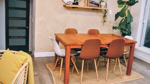 Ein Essbereich mit Holztisch und Stühlen, einem gewebten Teppich, einem Korbstuhl mit gelbem Kissen, einer großen Topfpflanze und einem Wandregal mit Dekoration.