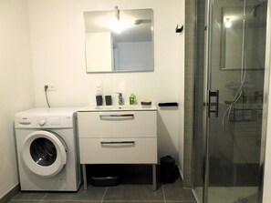 A compact bathroom with a washing machine, a sink with toiletries, and a glass shower enclosure. - Charming apartment💕 Near beaches and shops. Secure parking (Loctudy)