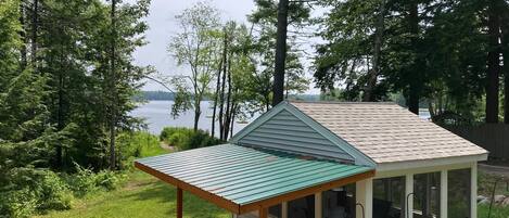 A small cabin with a covered porch, a deck, and a view of a lake surrounded by trees.