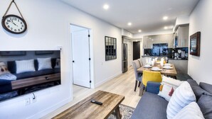 A modern living room with a sofa, a coffee table, and a TV. There is a dining area with a table and chairs, and a kitchen in the background. - Elegant 2BR Suite Close to Transit (Montréal)