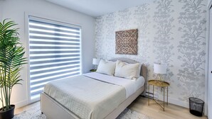A bedroom with a bed, a potted plant, a window with blinds, a wall with floral wallpaper, and a bedside table with a lamp. - Elegant 2BR Suite Close to Transit (Montréal)