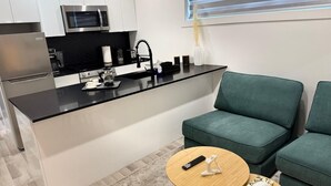 A compact kitchen with a black countertop, white cabinets, and a stainless steel refrigerator. There is a green sofa and a small round wooden table with a remote control and a decorative item. - NDG Treasure in Montreal! Value & Travel Adventure (Montréal)