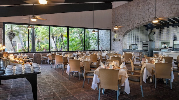 A restaurant with multiple tables set for dining, a kitchen area, and large windows offering a view of greenery. - El Cid Granada Hotel - All Inclusive (Mazatlán)