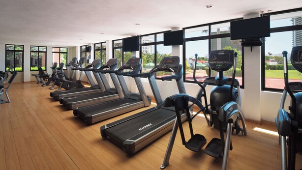 A gym with treadmills and ellipticals, large windows, and a view of greenery and buildings. - El Cid Granada Hotel - All Inclusive (Mazatlán)