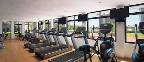 Un gimnasio con cintas de correr y elípticas, ventanas amplias y vistas a zonas verdes y edificios.