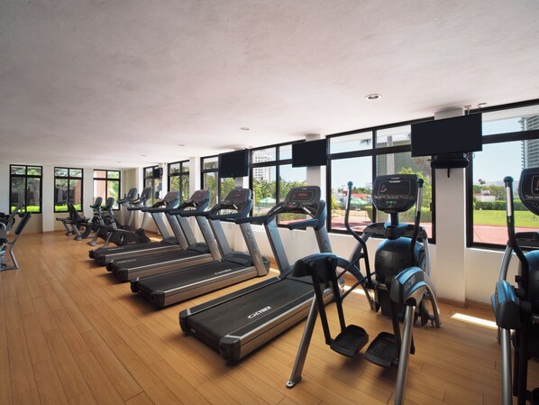 Ein Fitnessstudio mit Laufbändern und Crosstrainern, großen Fenstern und Blick auf Grünflächen und Gebäude.