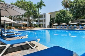 A swimming pool with lounge chairs and umbrellas, surrounded by palm trees and a building with multiple floors. - El Cid Granada Hotel - All Inclusive (Mazatlán)