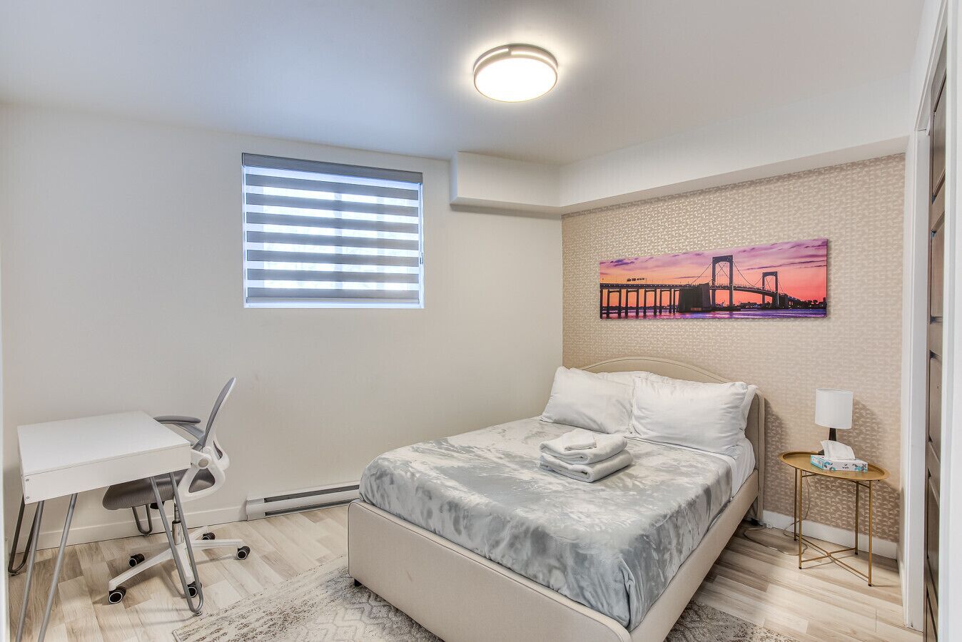 Un dormitorio con cama, escritorio, silla, ventana con persianas, un cuadro en la pared y una mesita de noche con lámpara.