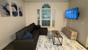 A living room with a sectional sofa, a coffee table, a TV, and a window with blinds. - LuxeWoodlands Villa: Cozy Stay (Conroe)