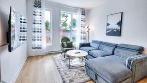 Ein modernes Wohnzimmer mit einem grauen Sofa, einem runden Couchtisch und einem grünen Sessel. Große Fenster mit durchsichtigen Vorhängen, ein flachbildfernseher an der Wand und ein gerahmtes Bild über dem Sofa.