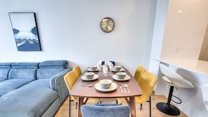 Un comedor moderno con una mesa de madera dispuesta para cuatro personas, rodeada de sillas amarillas y azules. Un reloj está colgado en la pared sobre la mesa.