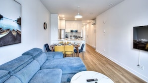 Una sala de estar moderna con un sofá azul, una mesa de comedor con sillas, una zona de cocina con armarios blancos y un televisor de pantalla plana montado en la pared.