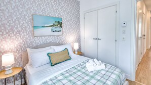 Un dormitorio ordenado con cama, almohadas, una lámpara de noche, un cuadro de una escena de playa y un armario.