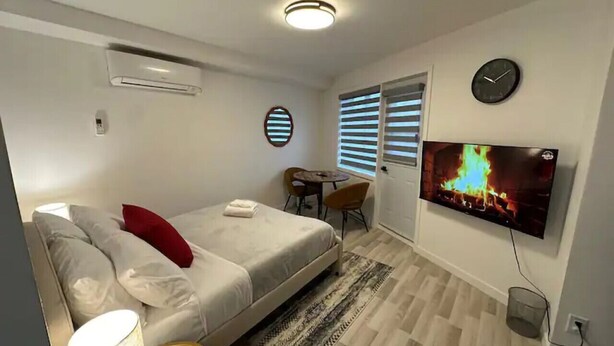 Quarto de hotel moderno com cama, televisão exibindo uma lareira, pequena mesa com cadeiras e relógio na parede.