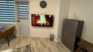 Uma sala de estar com uma televisão de tela plana na parede, um relógio, uma porta, uma mesa com uma cadeira e uma geladeira.