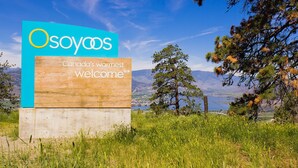 A sign with the text "Osoyacos" and "Canada's warmest welcome" against a scenic backdrop of mountains, trees, and a clear sky. - $2,175 / 3br - 1450ft2 - 3 Beds 2 Baths House (Osoyoos) (Osoyoos)