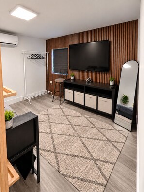 Een moderne woonkamer met een flatscreen televisie, een houten accentmuur, een zithoek met een krukje, een bijzettafeltje en een kamerkleed met een patroon.