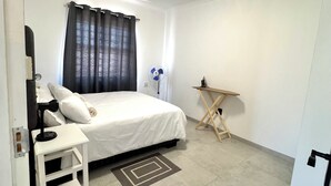 Un dormitorio con cama, mesita de noche, escritorio y ventilador de pie.