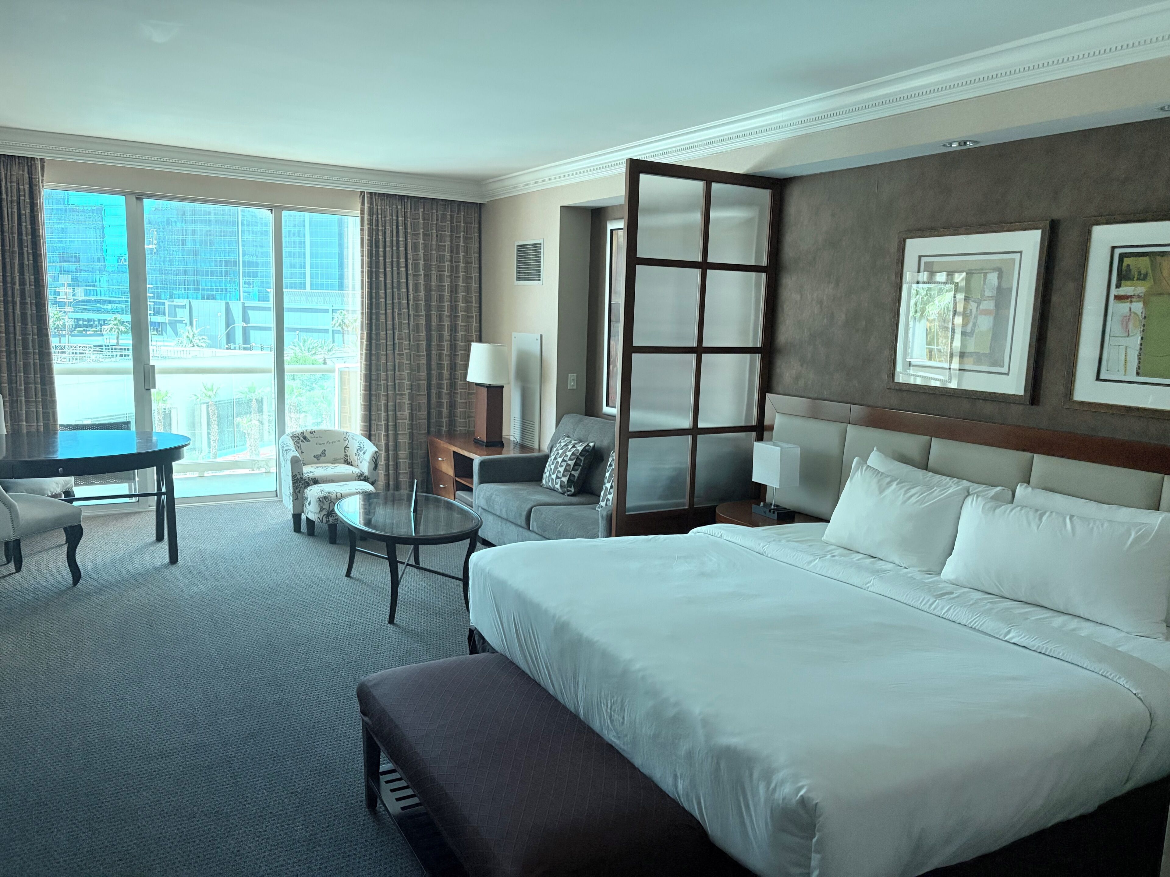 A hotel room with a large bed, a desk, a chair, a small table, and a view of the city.