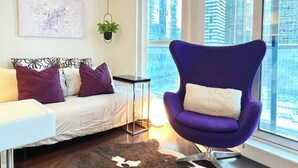 Ein modernes Schlafzimmer mit einem Bett, einem violetten Sessel, einem kleinen Tisch, einer Hängepflanze und einem Blick auf die Stadt durch große Fenster.