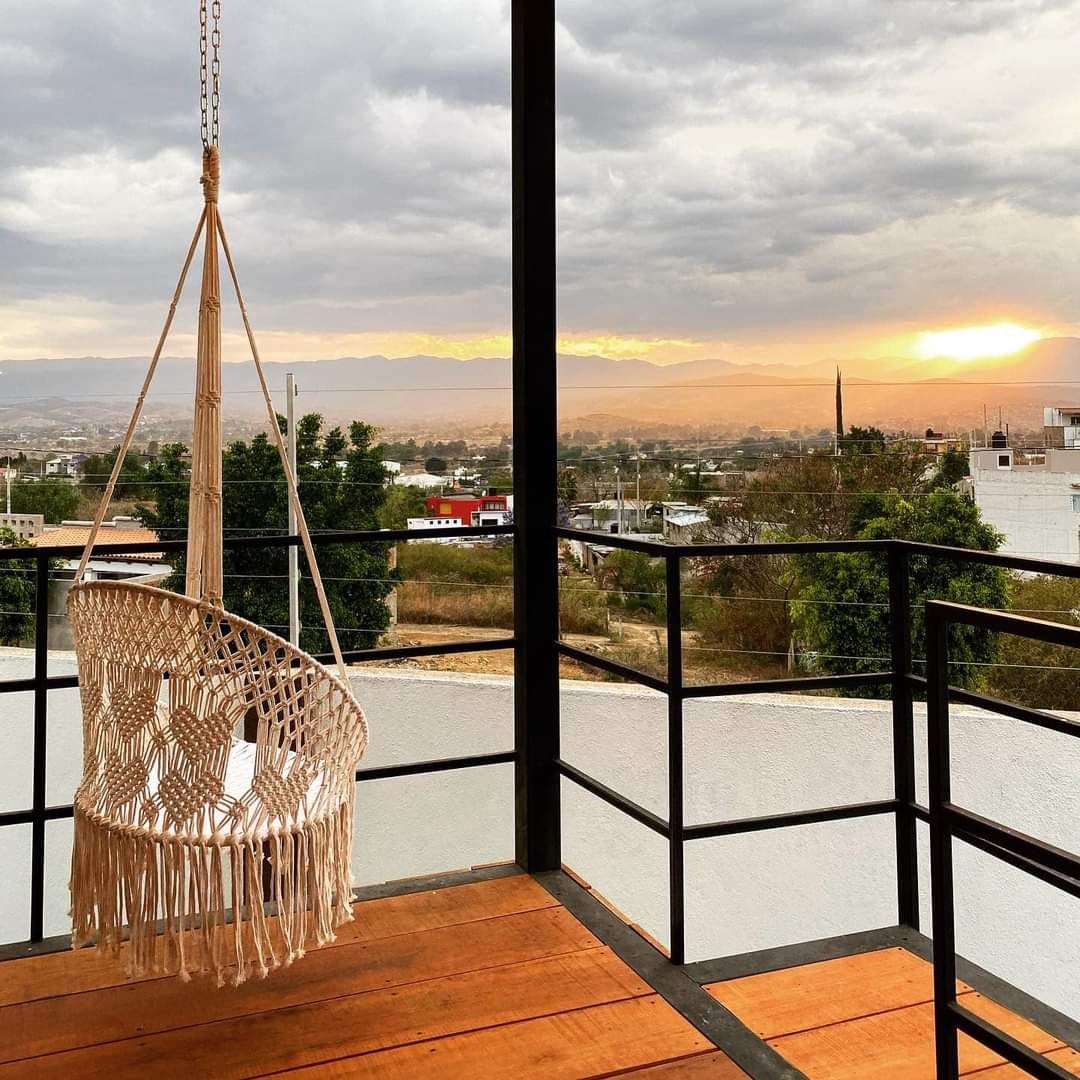 Una habitación con una silla colgante de macramé, vista a un paisaje urbano y una puesta de sol.