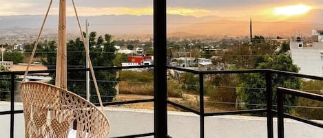 Una habitación con una silla colgante de macramé, vista a un paisaje urbano y una puesta de sol.