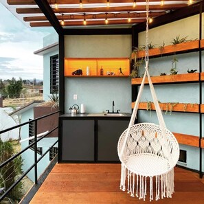 Un balcon agrémenté d’un fauteuil suspendu en macramé, d’un petit coin cuisine et d’étagères où sont disposées des plantes.