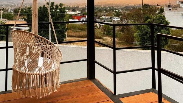A room with a hanging macrame chair, a view of a cityscape, and a sunset. - Casa Terraza Oaxaca (Oaxaca de Juárez)