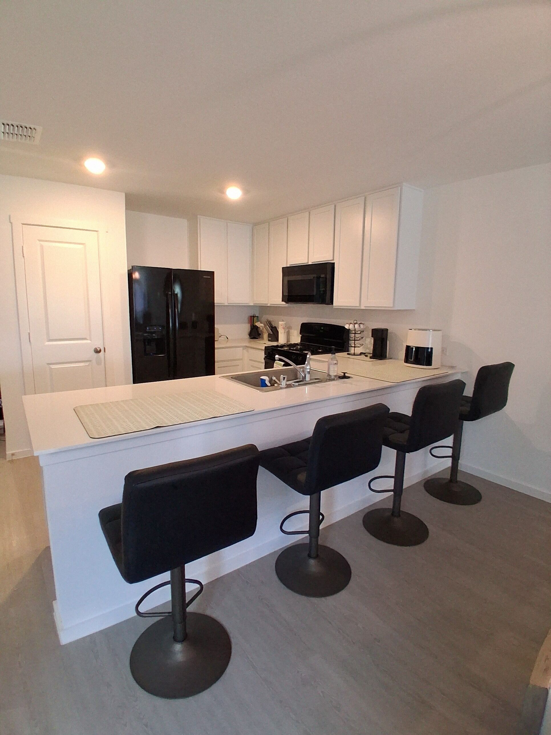 A modern kitchen with white cabinetry, a black refrigerator, a microwave, and a gas stove. There are four black bar stools around the island.