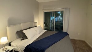 A bedroom with a bed, a nightstand, a lamp, and a sliding glass door leading to a balcony. - Chic Apt in The Woodlands-Parking-WI-FI-King Bed (Conroe)