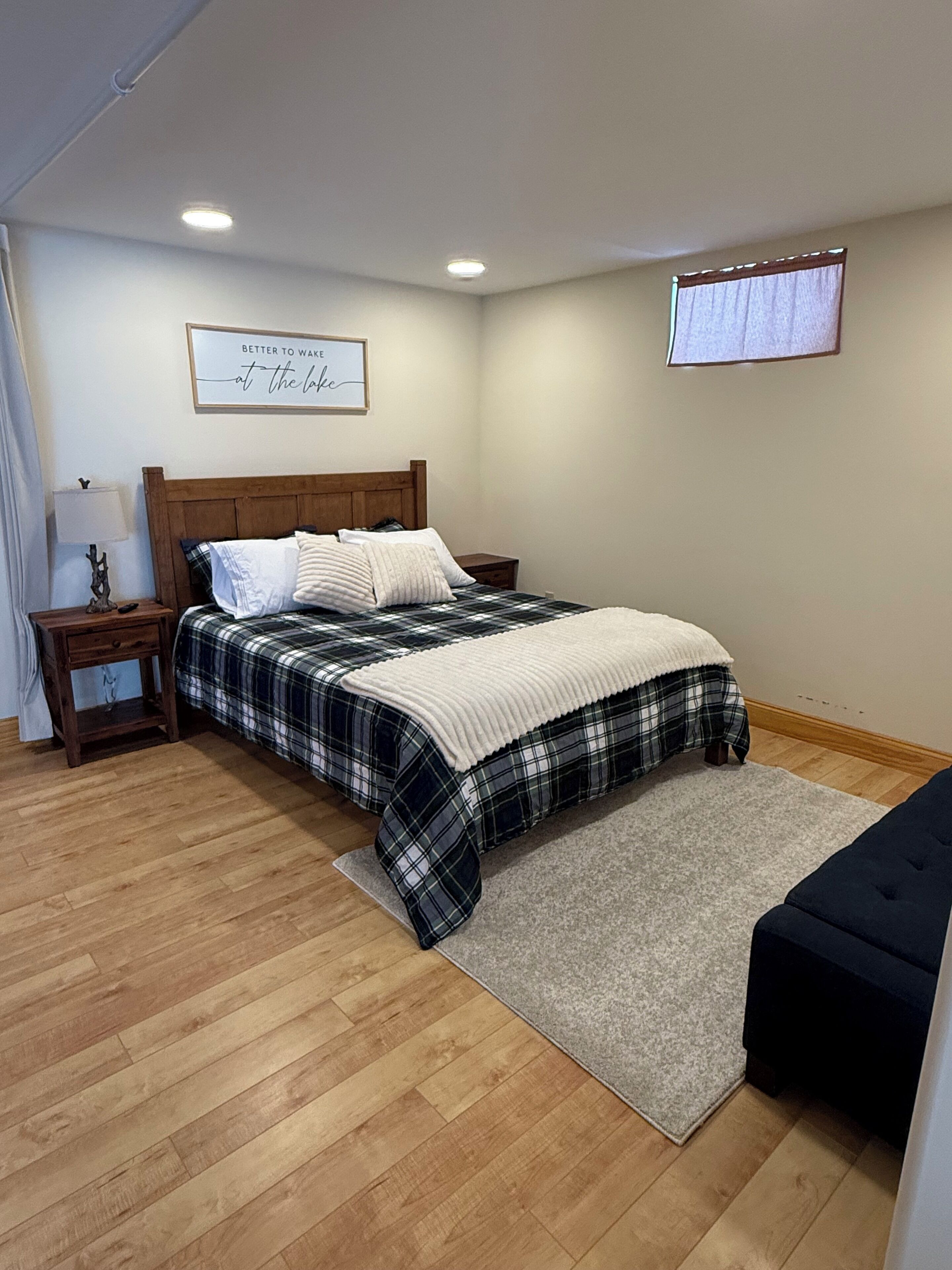 Une chambre à coucher bien rangée, avec un lit en bois, une table de chevet sur laquelle se trouve une lampe, un petit tapis et, au mur, une citation inspirante encadrée.