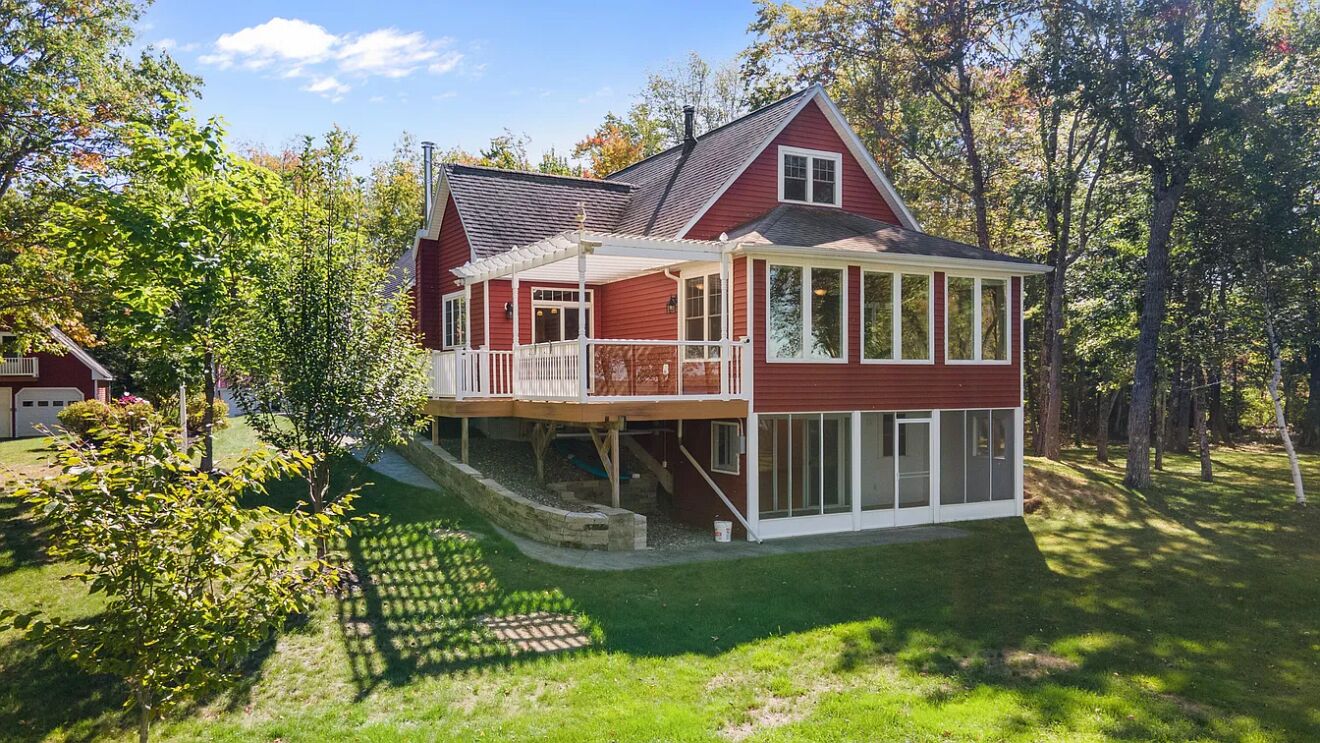 Ein zweistöckiges Haus mit einer überdachten Veranda und einer kleinen Terrasse, umgeben von Bäumen und einem gepflegten Rasen.